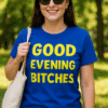 Vidám nő napsütéses parkban, kék pólón "Good Evening Bitches" felirattal, nyári szabadidő ruhában.