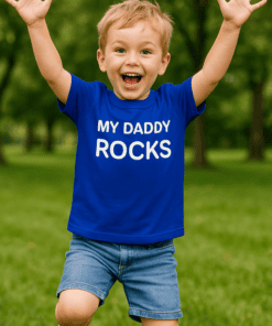 Vidám kisfiú játszik a szabadban, kék pólóban, "My Daddy Rocks" felirattal.