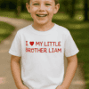 Gyermek mosolyogva áll a természetben, fehér pólón, amelyen a "I love my little brother Liam" felira.