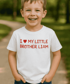 Gyermek mosolyogva áll a természetben, fehér pólón, amelyen a "I love my little brother Liam" felira.
