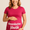 Bézs háttér előtt álló mosolygó nő, aki egy pink pólón viseli a "Novemberben jövök" feliratot.