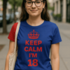 Fiatal nő az utcán, "Keep Calm I'm 18" feliratos kék pólóban, városi környezetben.