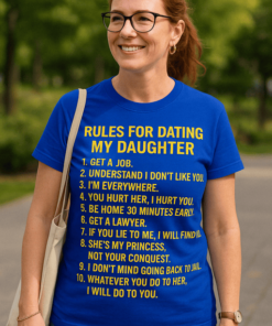 Vidám nő kék pólóban, humoros szöveggel, parkban, nyári napfényben.